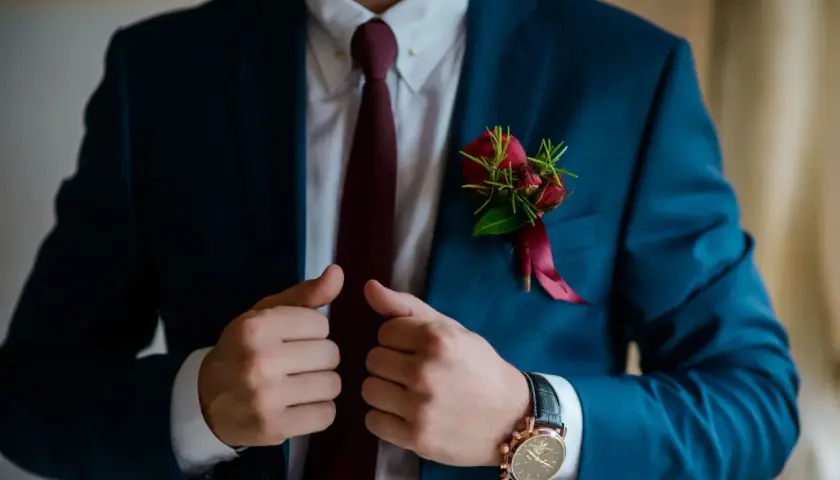 Elegancki mężczyzna w granatowym garniturze, białej koszuli i bordowym krawacie. Na klapie marynarki ozdobna butonierka z czerwonych róż.
