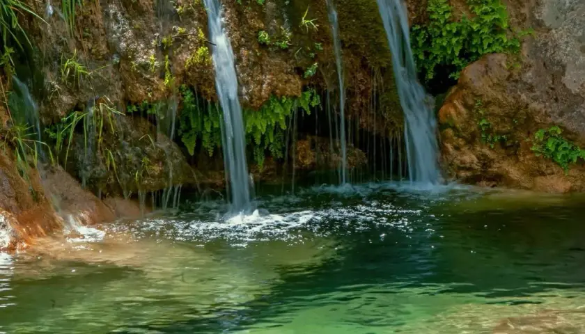 Cascada de la Osera, con agua cayendo sobre rocas cubiertas de musgo y vegetación exuberante, formando un estanque de aguas cristalinas.