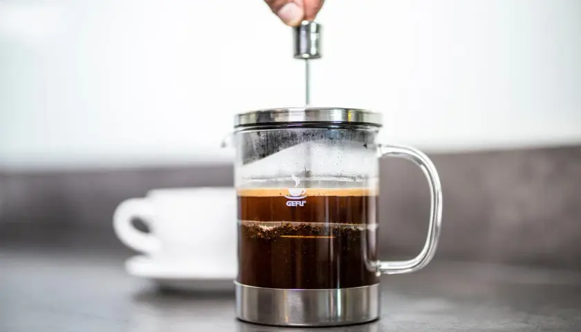 Eine Hand drückt den Stempel herunter, um Kaffee im Kaffeebereiter zuzubereiten. Daneben steht eine weiße Tasse.