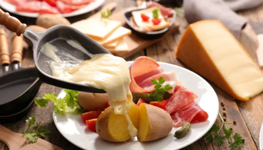 Roztopiony ser raclette spływa na ziemniaki i wędliny. Obok kawałek sera i talerz z dodatkami.