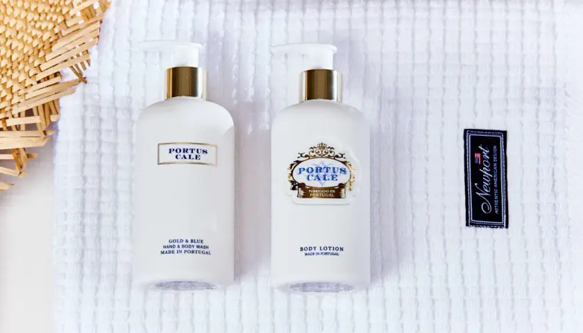 Two white pump bottles of Portus Cale body wash and lotion on a white textured towel, with a woven straw fan and a Newport tag.