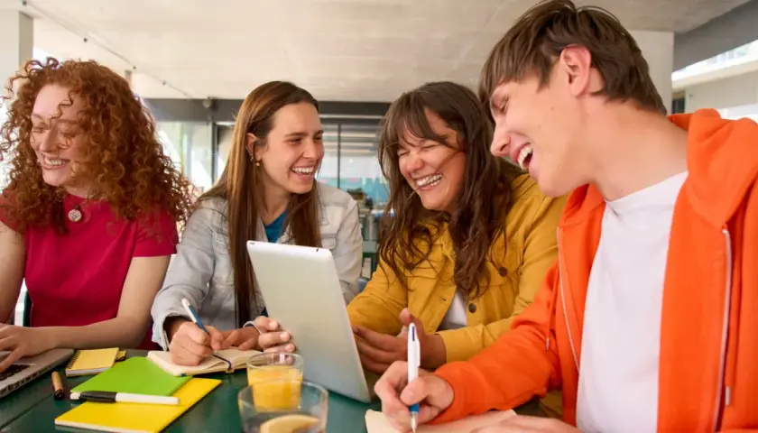 Uśmiechnięci studenci, którzy uczą się psychologii na studiach zaocznych, pracują razem przy stole, korzystając z laptopa i tabletu.