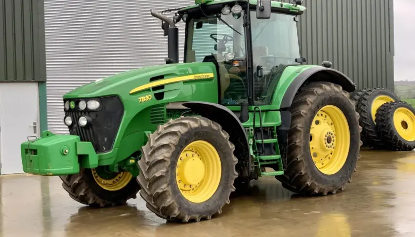 Zielony John Deere 7830 z żółtymi felgami stoi na mokrej posadzce przed budynkiem gospodarczym.