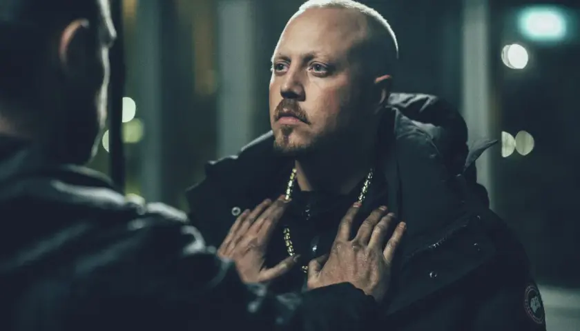 En dramatisk scen från en sebbe stakset film. Två män möts i mörkret, en med guldhalsband, den andra med händerna på hans bröst.
