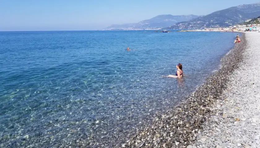Une femme se baigne dans les eaux claires de la plage de galets de Ventimiglia, avec des montagnes en arrière-plan.