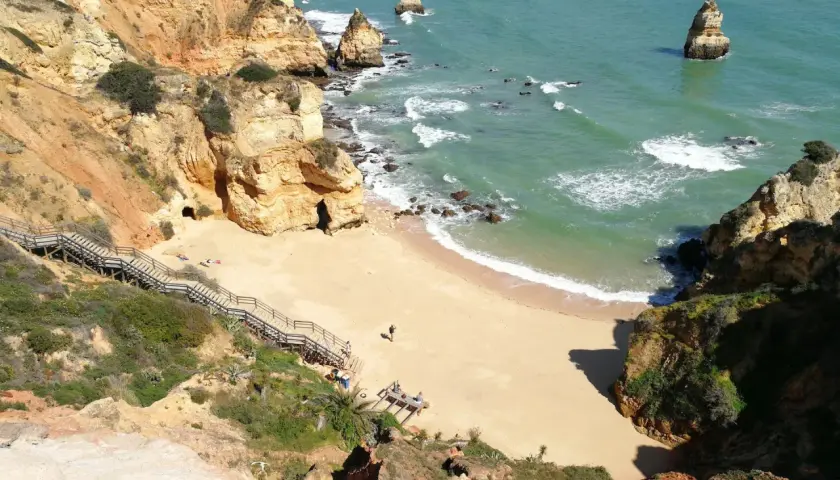Acantilados dorados y mar turquesa en las **islas de Portugal**. Escaleras de madera descienden a una playa de arena dorada, con formaciones rocosas emergiendo del agua.