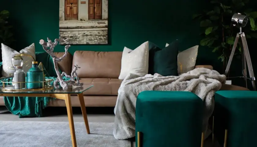 Elegancki salon z butelkową zielenią na ścianach. Brązowa kanapa, złote i zielone poduszki, puszysty koc. Stolik kawowy ze szklanym blatem i drewnianymi nogami.