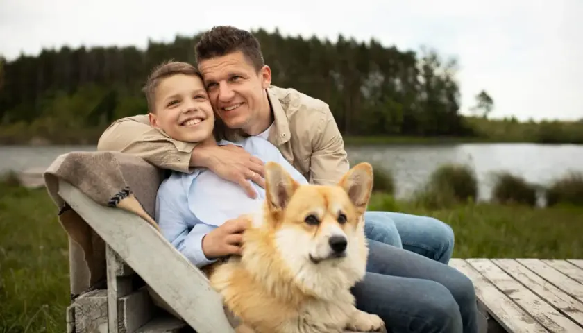 Ojciec i syn siedzą przy jeziorze z psem. Corgi kładzie się na kolanach chłopca, bo czuje się bezpiecznie i kochany.