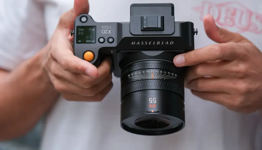 Dłonie trzymają aparat Hasselblad X2D 100C z obiektywem XCD 55mm. Widoczne detale aparatu i obiektywu.