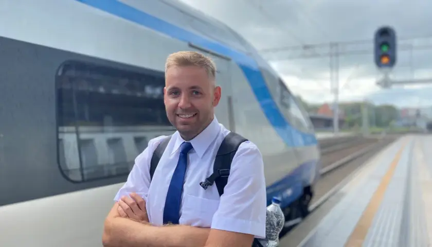 Uśmiechnięty mężczyzna w białej koszuli i niebieskim krawacie, gotowy do pracy. To on prowadzi pociąg, który stoi obok na torach.