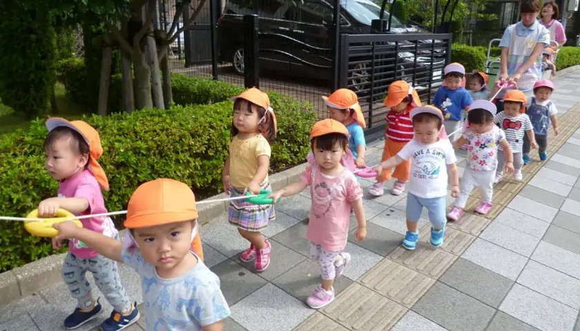 Un gruppo di bambini in Giappone, con cappellini arancioni, cammina in fila tenendo una corda.