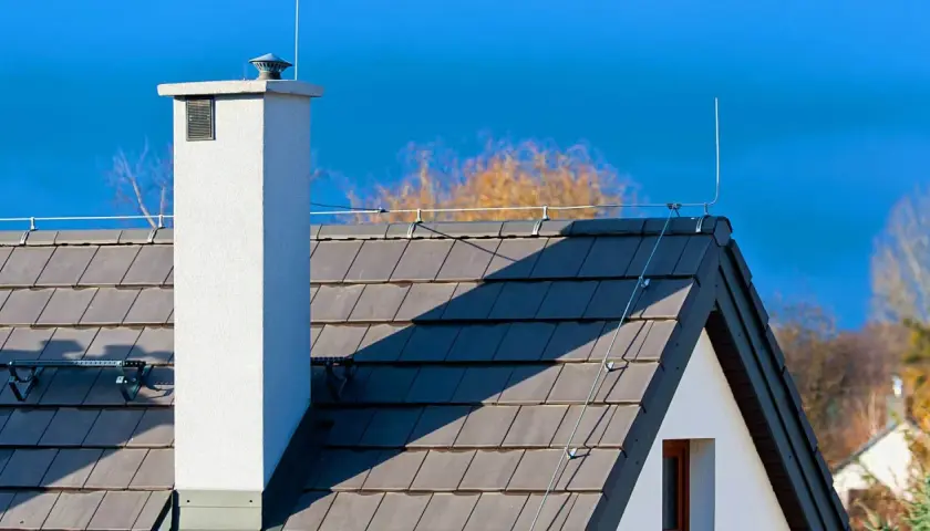 Biały komin i dachówka na tle błękitnego nieba. Na dachu widoczny jest piorunochron, chroniący dom przed burzą.