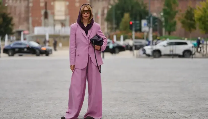 Kobieta w garniturze w kolorze mauve, z szerokimi nogawkami spodni, pozuje na tle miasta.