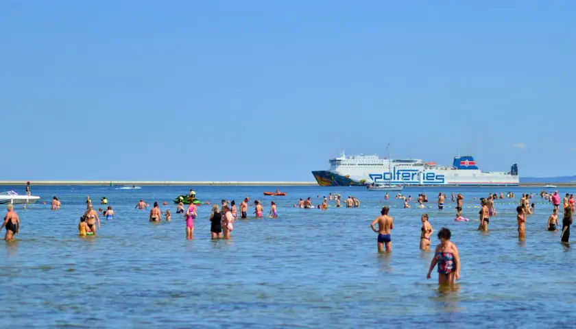Ludzie kąpią się w morzu, a w tle widać prom Polferries. Szukasz informacji o prom Świnoujście-Ystad cena?