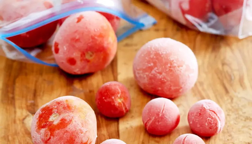 Gefrorene Tomaten liegen auf einem Holzbrett. Einige sind in Gefrierbeuteln, andere einzeln. Perfekt für Soßen und Suppen, wenn man Tomaten einfrieren möchte.