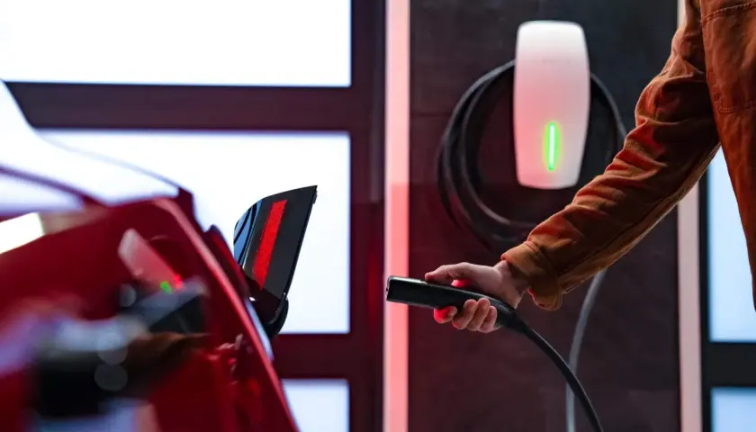 Ręka trzymająca ładowarkę do samochodu elektrycznego przy czerwonym aucie. Obok widać biały wallbox z zielonym wskaźnikiem.