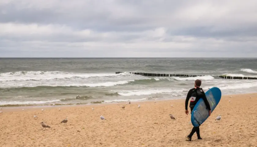 Surfer z deską na plaży w Juracie, szuka fal. W tle morze i mewy. Jedna z atrakcji nad polskim morzem.
