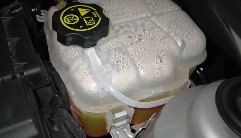 Depósito de expansión del coche con agua y burbujas. El tapón negro tiene símbolos de advertencia.