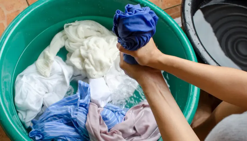 Mains lavant du linge dans une bassine verte. Un vêtement décoloré par le soleil, blanc et bleu, baigne dans l'eau.