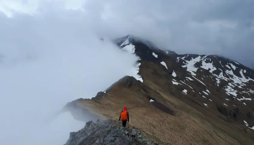 Trekking w Pirenejach. Samotny turysta w pomarańczowym plecaku idzie skalistą granią, otoczony mgłą i ośnieżonymi szczytami.