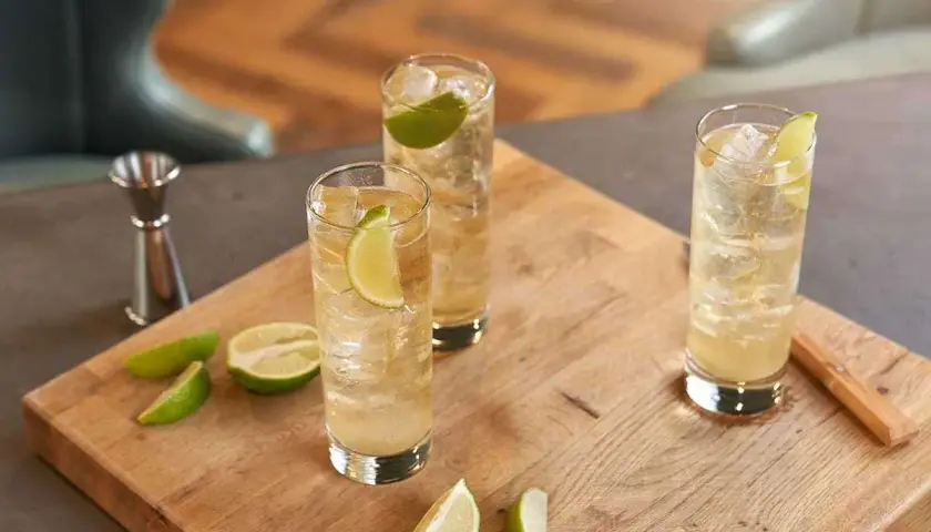 Trzy orzeźwiające whisky ze spritem, udekorowane plasterkami limonki, stoją na drewnianej desce. Obok leżą świeże kawałki limonki.