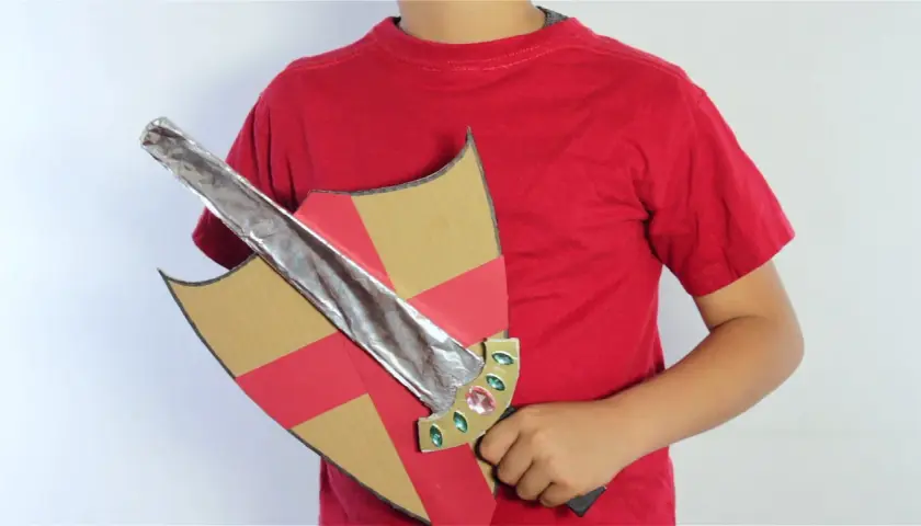 Un niño con camiseta roja sostiene un escudo de cartón con detalles rojos y dorados y una espada de cartón plateada, lista para la aventura.