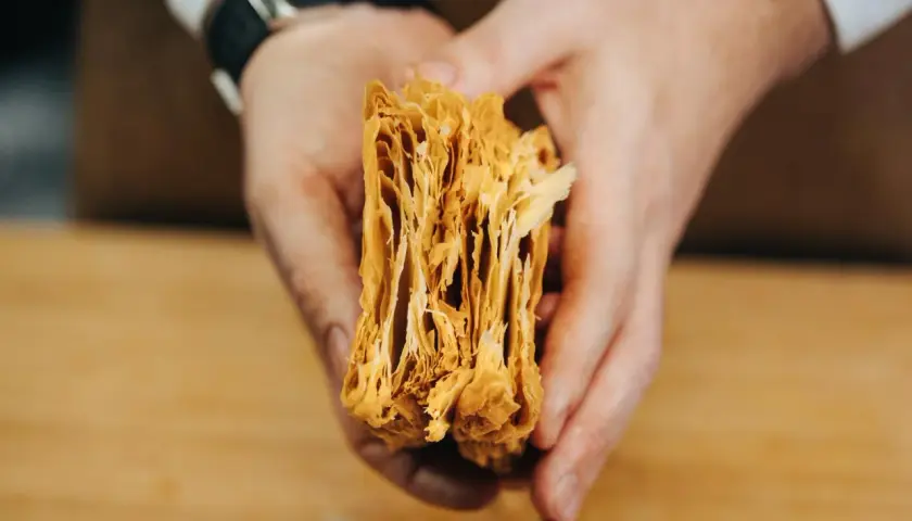 Manos sostienen un trozo de masa de hojaldre casera, mostrando sus finas y crujientes capas doradas.