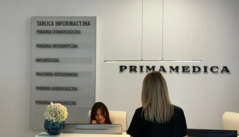 Recepcja kliniki PRIMAMEDICA z tablicą informacyjną i pracownicą.