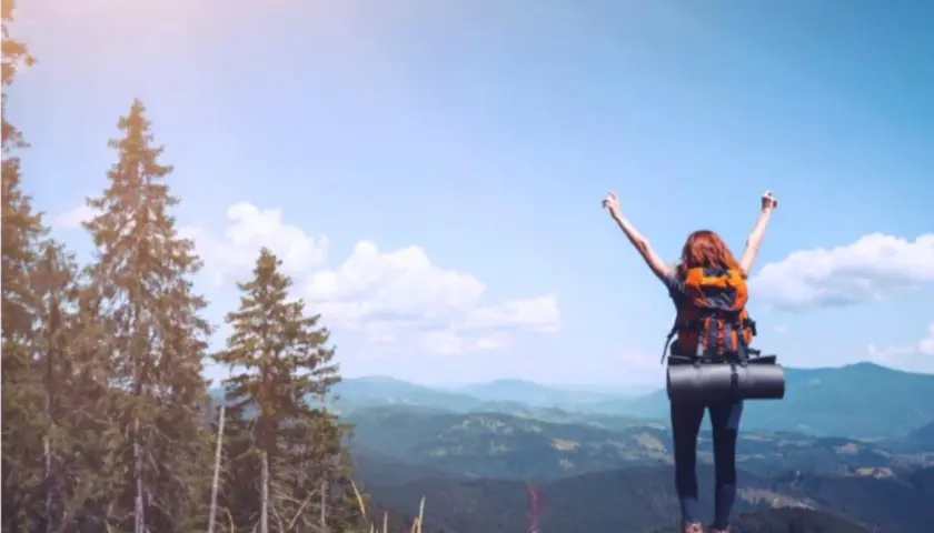 Donna con zaino in cima a una montagna, braccia alzate, esprime il bisogno di viaggiare psicologia.