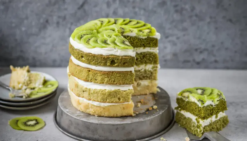 Eine mehrstöckige Matcha Torte mit Kiwi-Scheiben und Sahne. Ein Stück wurde bereits auf einem Teller serviert.