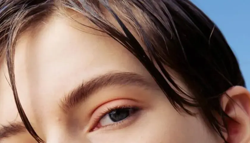 Primer plano de un ojo y cabello oscuro y mojado sobre fondo azul claro.