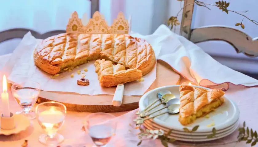 Une galette des rois dorée, prête à être partagée, avec une couronne posée à côté. Une part est déjà servie, invitant à la fête.