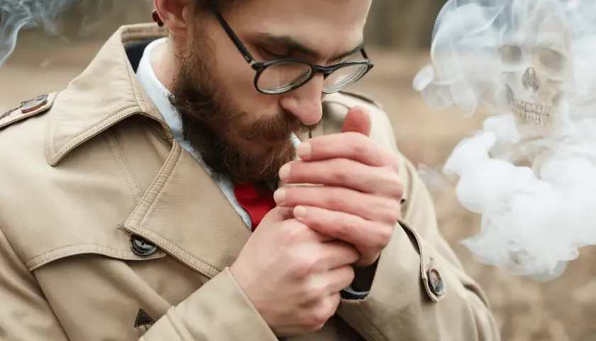 Hombre con barba enciende cigarrillo. El humo forma una calavera, advirtiendo sobre la alergia al humo del tabaco.