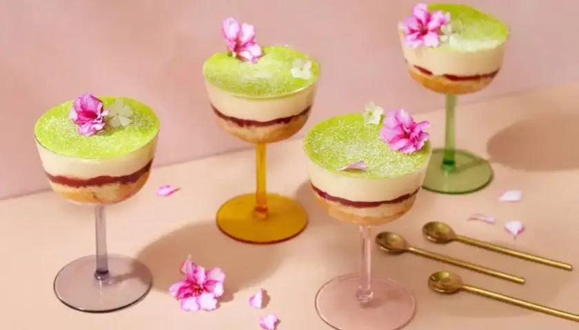 Vackra dessert i glas med lager av kaka, sylt och grön kräm, toppad med blommor. Guldfärgade skedar ligger bredvid.