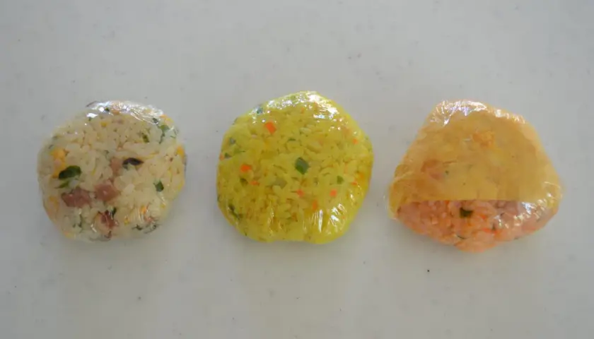 Three plastic-wrapped onigiri sit in a row. The left is white rice with green and brown bits. The middle is yellow rice with orange and green flecks. The right is orange rice with a yellow wrapper. Can you freeze onigiri?