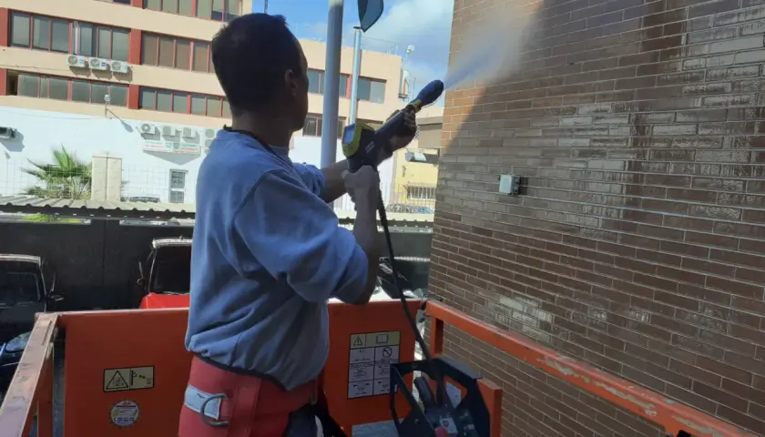 Hombre en plataforma elevadora realiza limpieza de fachadas con agua a presión, dejando la pared de ladrillo brillante y reluciente.