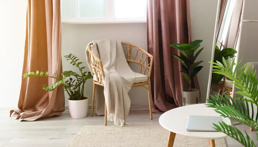 A cozy reading nook with a wicker chair draped in a blanket, flanked by pooled curtains in soft peach and dusty rose hues.