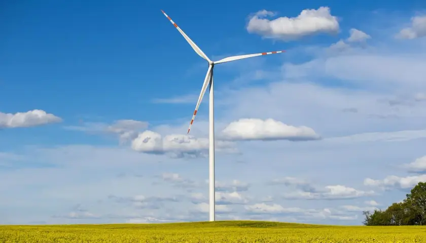 Wiatrak na tle błękitnego nieba z chmurami, pośród kwitnącego na żółto pola. To obraz branży OZE, co to jest energia odnawialna.