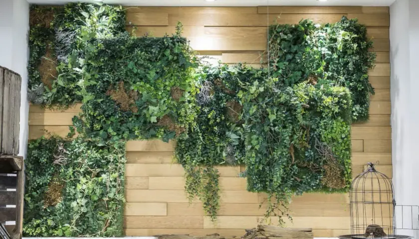 Eine üppige, grüne Pflanzenwand, die man leicht selber machen kann, schmückt eine Holzvertäfelung. Deko-Vogelkäfig und Treibholz runden das Bild ab.