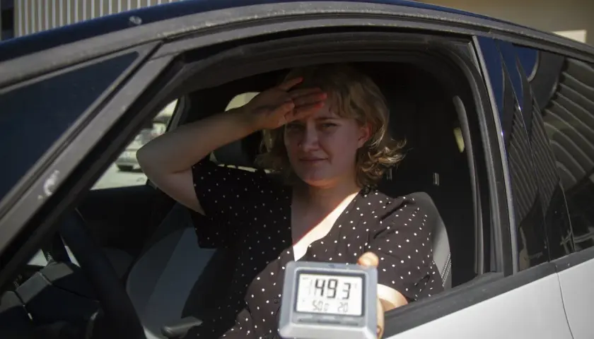 Kvinna i bil svettas och håller upp en termometer som visar 49,3 grader. Det är så varmt att bilen blir inte varm.