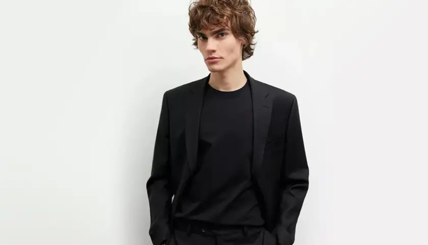 Młody mężczyzna w czarnym garniturze i czarnej koszuli, z rękami w kieszeniach, prezentuje elegancki styl.