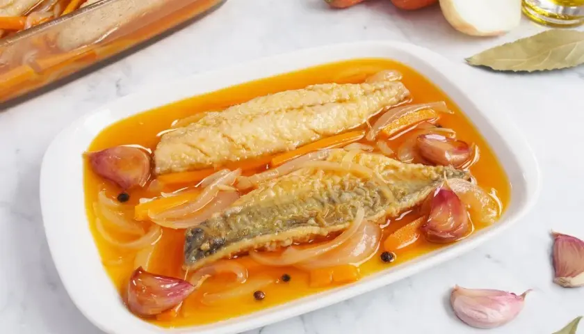 Dos filetes de caballa en escabeche, con zanahoria, cebolla y ajos, en una bandeja blanca.