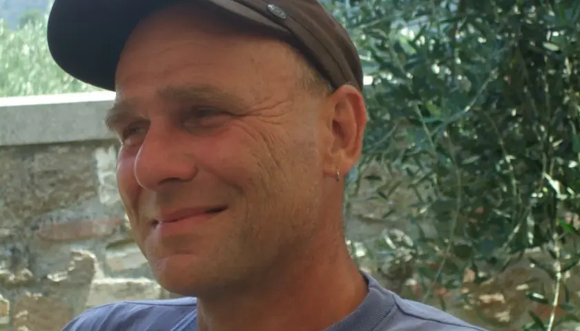 Martin Klein, Autor, lächelt in die Kamera, trägt eine braune Kappe und ein blaues T-Shirt. Im Hintergrund Olivenbäume.