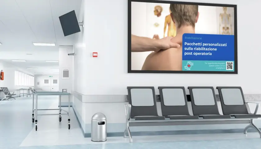 Corridoio di una sala di attesa con sedie, lettini e un monitor che pubblicizza pacchetti di riabilitazione post operatoria.