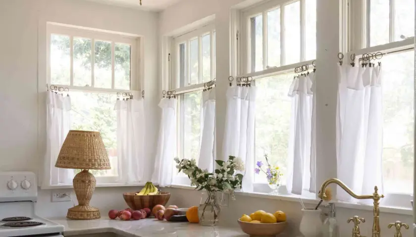 A bright kitchen with white diy cafe curtains framing the windows. A wicker lamp, fruit bowls, and a brass faucet add charm.