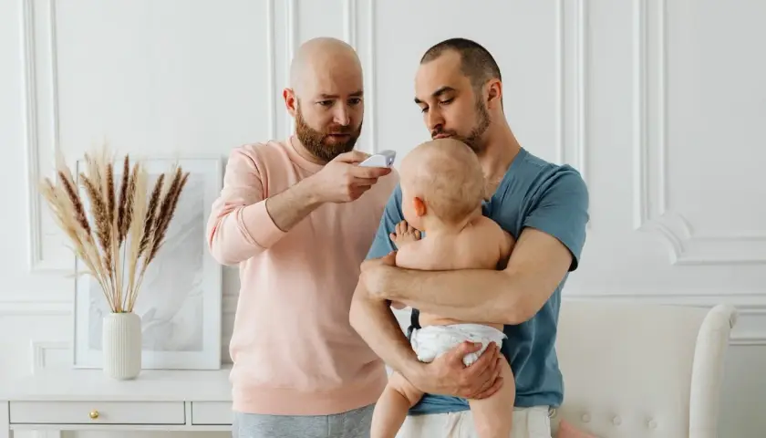 Två pappor kollar febern på sitt barn. En pappa håller barnet, den andra mäter temperaturen. Frågan "hur får man bort snuva" kan dyka upp när man tar hand om små barn.