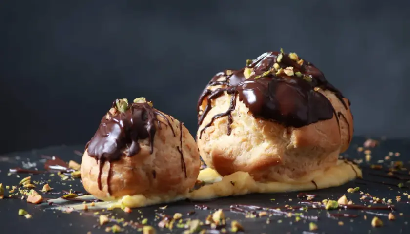 Deux profiteroles au chocolat, garnis de crème pâtissière et saupoudrés de pistaches concassées, sur un fond sombre.