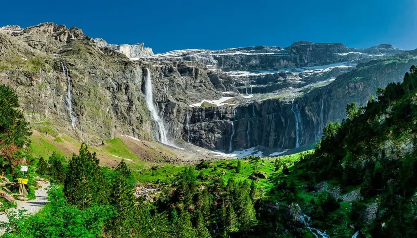 Cascadas impresionantes caen por acantilados rocosos cerca del pueblo de Gavarnie. Un sendero serpentea entre árboles verdes y prados exuberantes.