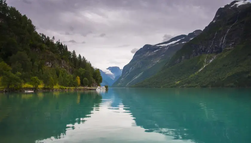 Turkusowa woda odbija skaliste góry i lasy. Statek cumuje przy brzegu, w oddali widać lodowiec. Piękne fiordy w Norwegii.