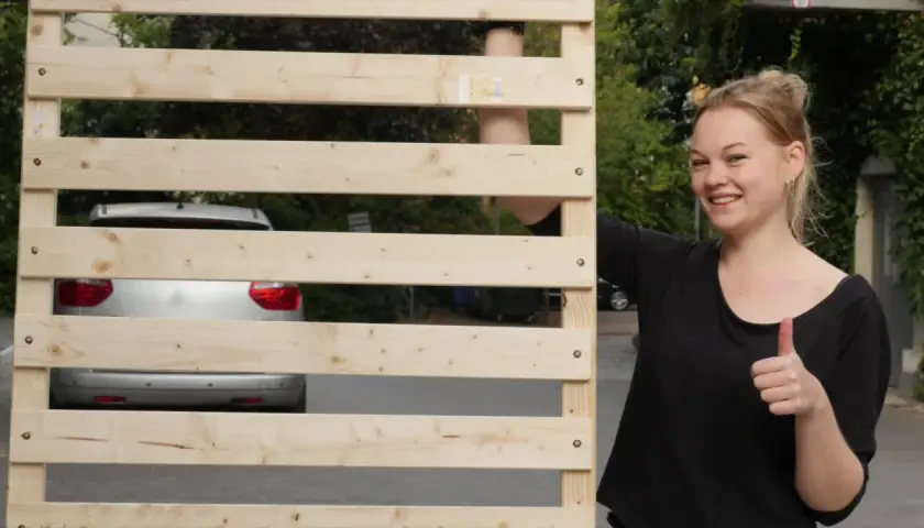 Junge Frau zeigt Daumen hoch neben selbstgebautem Lattenrost. Ein Auto ist im Hintergrund zu sehen.